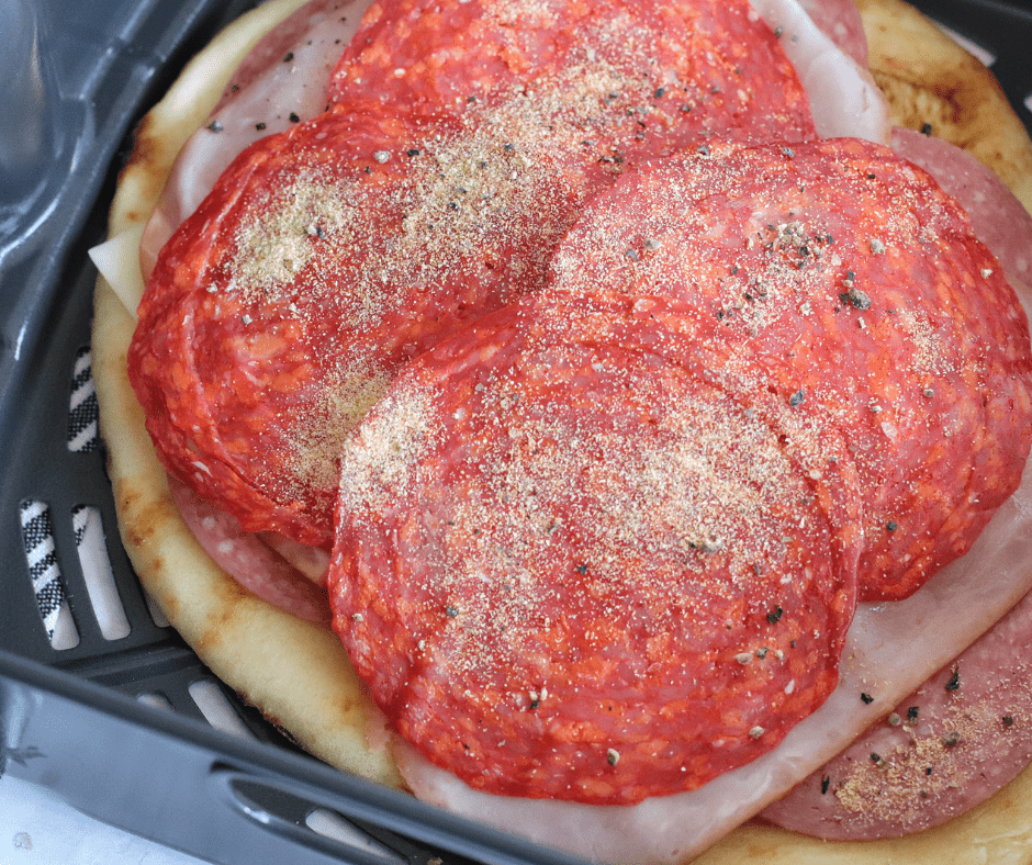 Flatbread topped with cheese, salami, ham, pepperoni, pepperoncini, and seasonings ready for air frying.