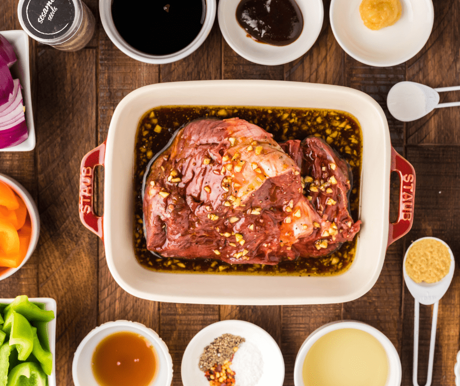 Top view of a dish with beef steak in it, marinating in a brown sauce. There are small white bowls and spoons filled with other ingredients around the edge of the frame.