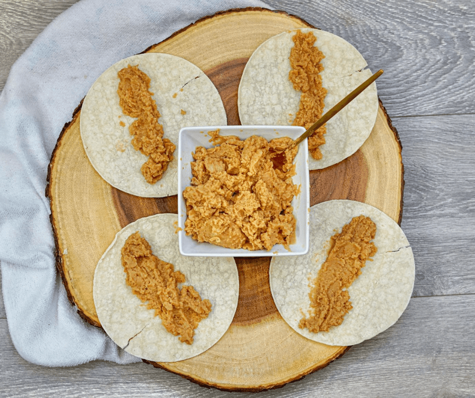 Refried beans and shredded cheddar spread down the center of a corn tortilla