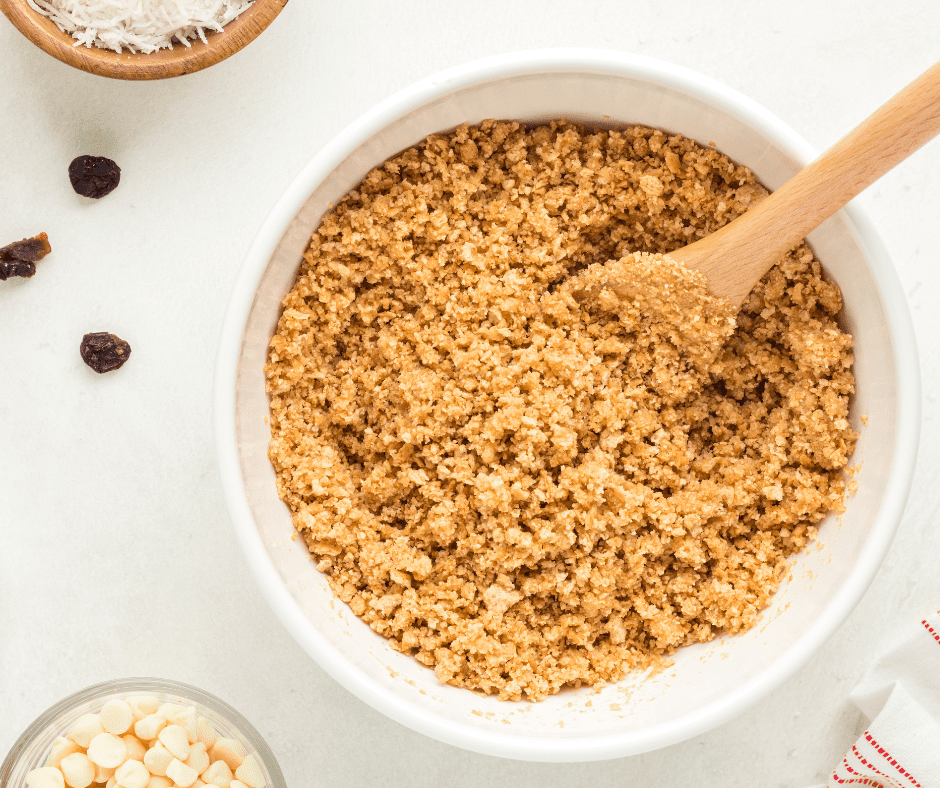 A medium glass bowl filled with a mixture of golden graham cracker crumbs and melted butter being stirred together with a spatula.