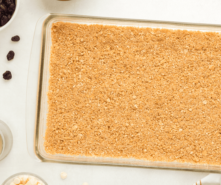 Hands firmly pressing a layer of buttery graham cracker crumbs into the bottom of a greased square baking pan to create a smooth, even cookie bar base.
