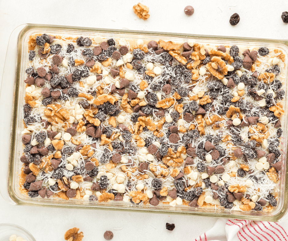 A stream of creamy sweetened condensed milk drizzled over a graham cracker crust, topped with a colorful layer of dried cranberries, chopped walnuts, shredded coconut, and chocolate chips.