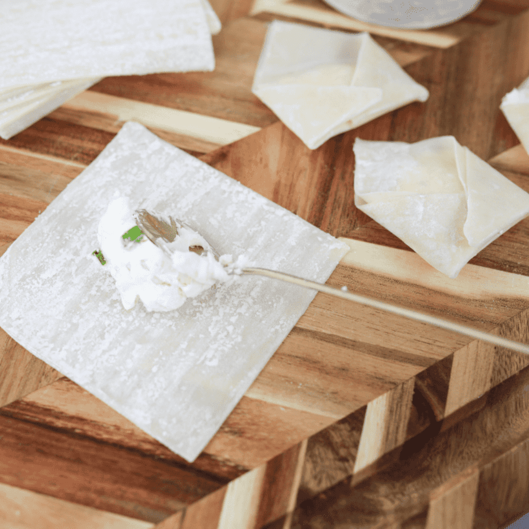 A wonton wrapper on a flat surface with a teaspoon of cream cheese mixture placed in the center, ready for folding.