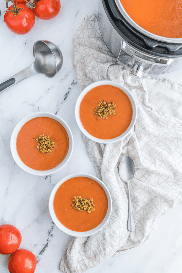 A steaming white bowl of creamy tomato soup garnished with fresh basil leaves and crispy croutons, served next to a grilled cheese sandwich on a wooden table.