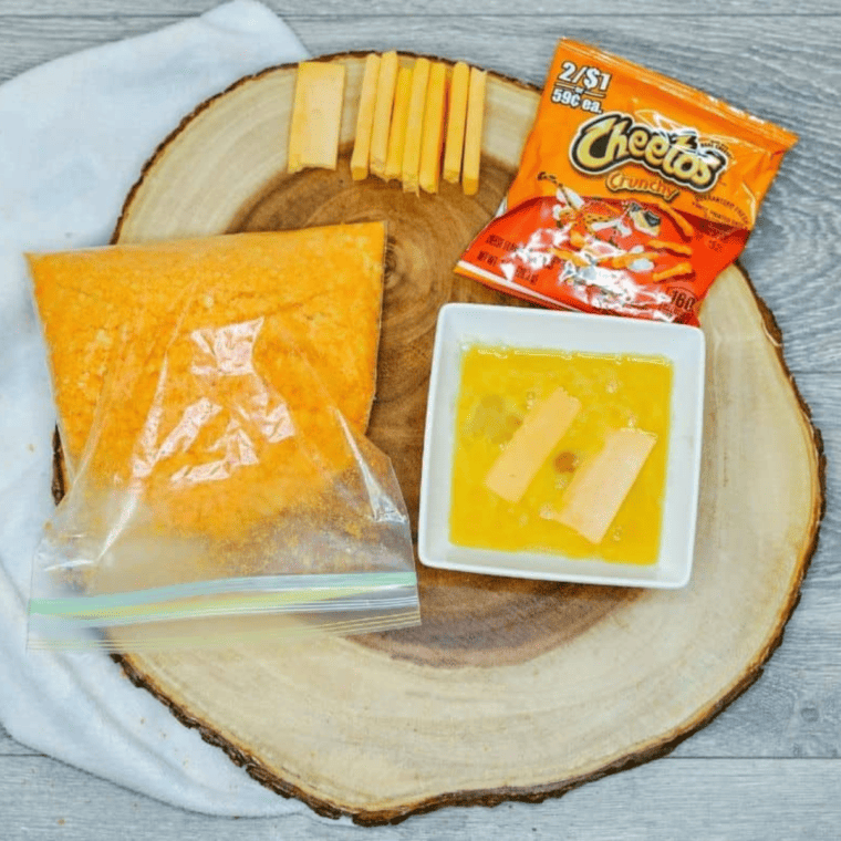 Three shallow bowls set up for breading: flour with garlic powder, beaten eggs, and crushed Cheetos for coating mozzarella sticks.