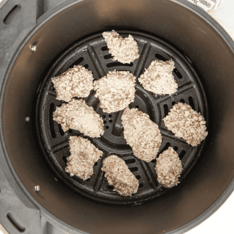 Air fryer basket filled with popcorn chicken in a single layer being sprayed with oil before crisping at high heat