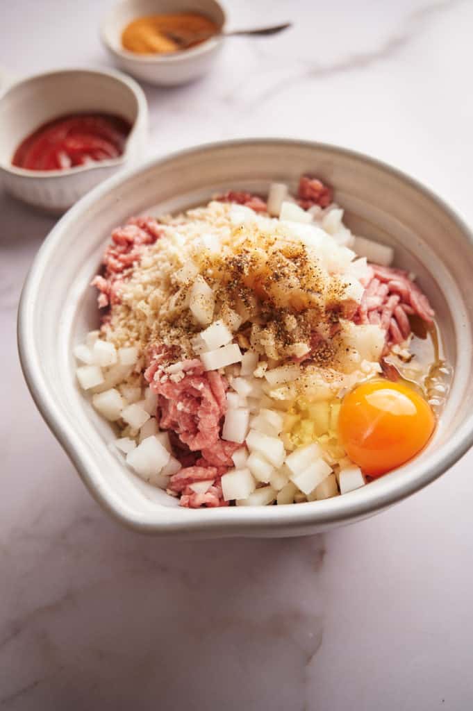 Mixing ground beef, chopped onions, bourbon, garlic, egg, Worcestershire sauce, steak seasoning, milk, and breadcrumbs in a large bowl for meatloaf preparation.