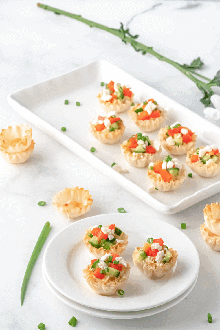 A platter of crispy Greek Hummus Phyllo Bites topped with diced cucumbers, cherry tomatoes, olives, and crumbled feta cheese.