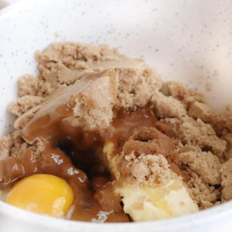 A mixing bowl filled with creamy dough as butter, brown sugar, vanilla extract, and egg are blended together, with flour, salt, and baking powder gradually added, and chocolate chips folded in before refrigeration.