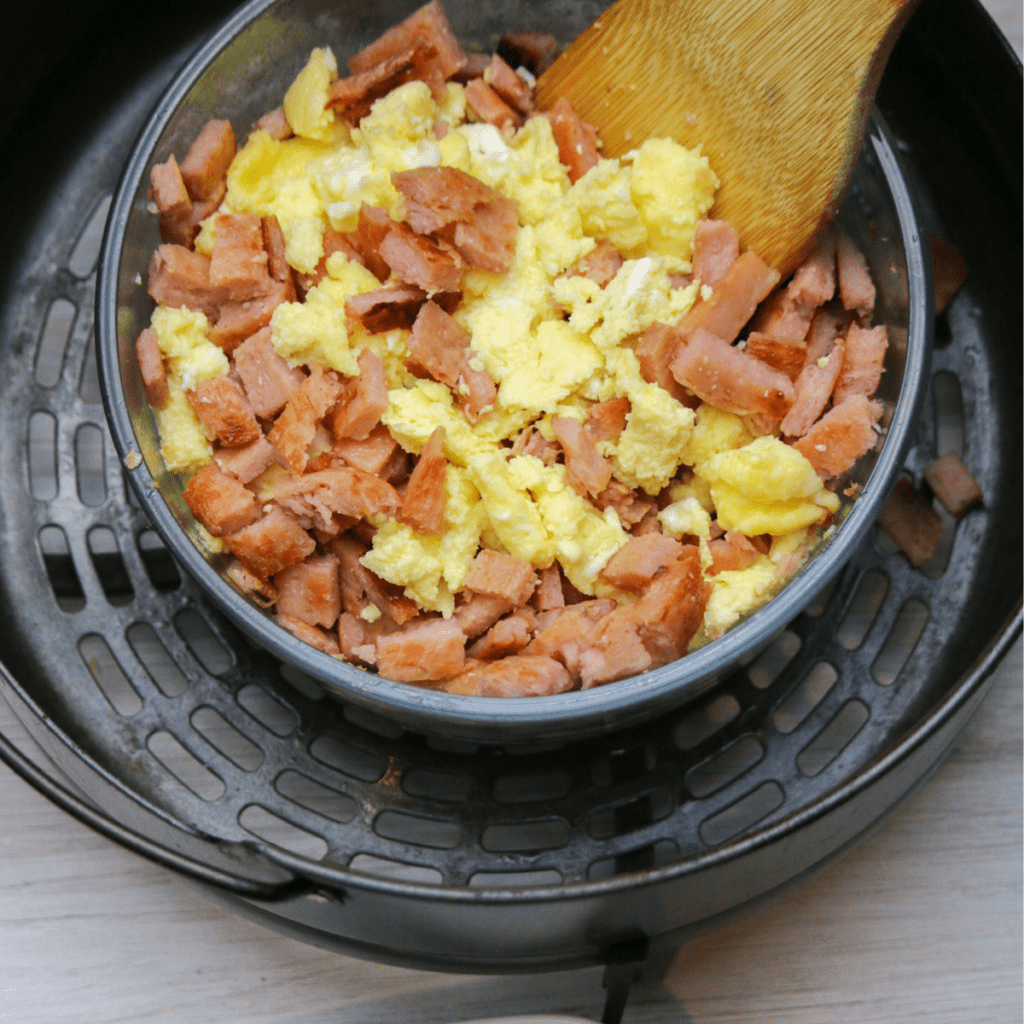 Scrambled eggs cooking in air fryer with butter and cream, stirred halfway through for soft texture.