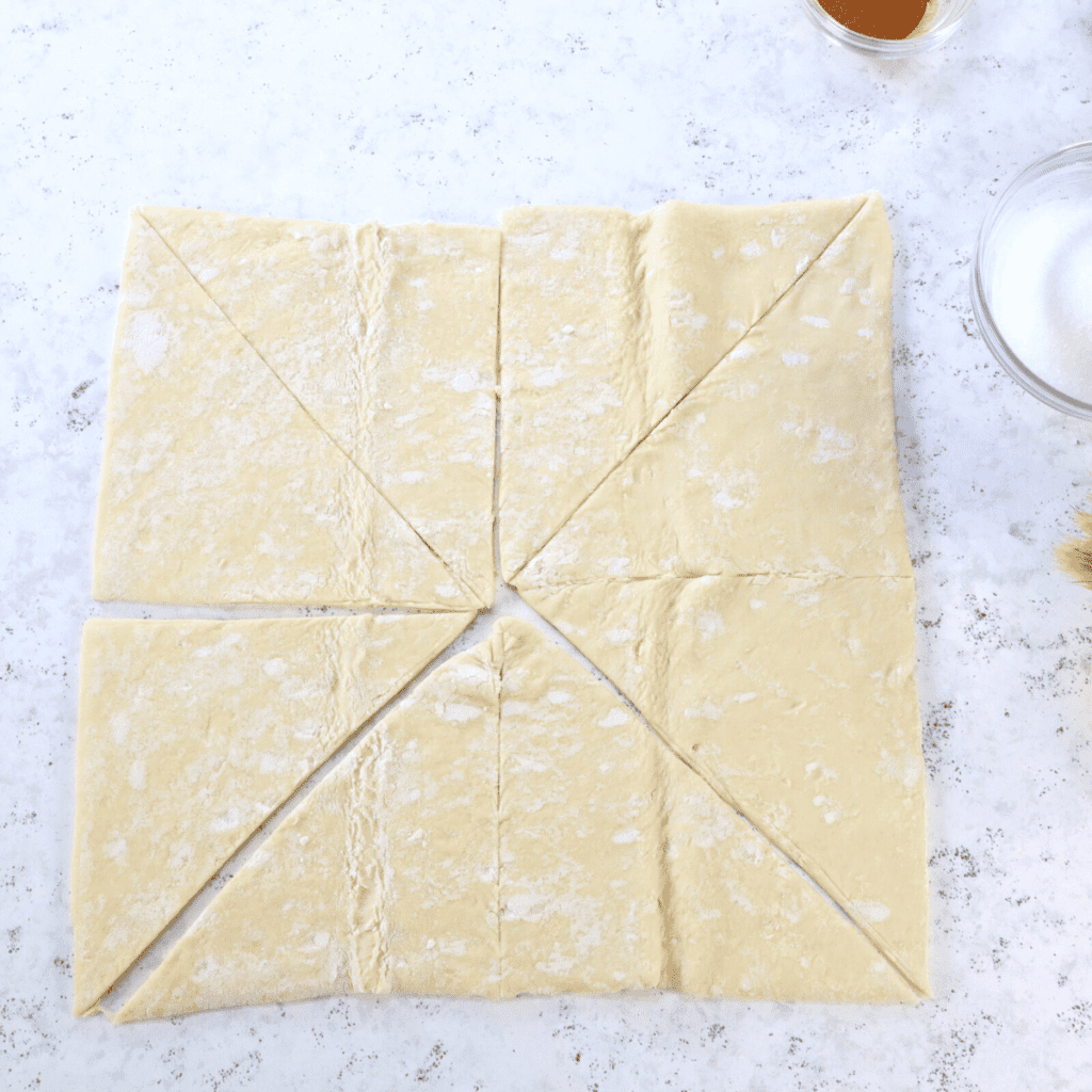Cutting dough on a floured surface into squares, then slicing into triangles using a pizza cutter for churro preparation.
