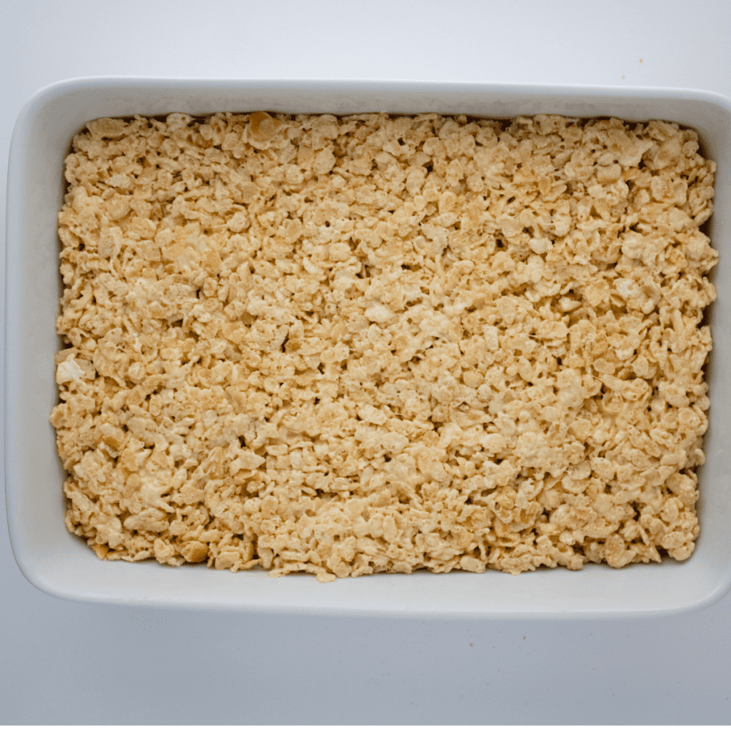 Pouring the banana pudding Rice Krispie mixture into a greased dish, drizzling melted white chocolate on top, and garnishing with extra vanilla wafers.