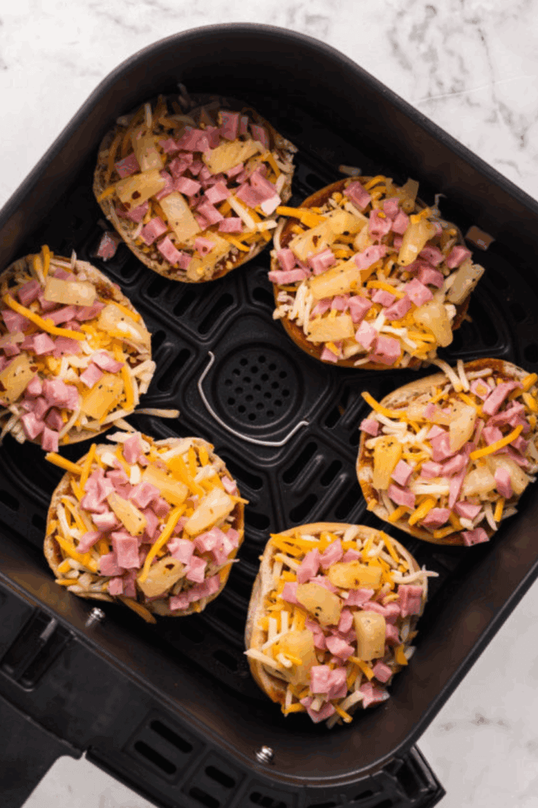 Bagel halves topped with BBQ sauce, cheese, ham, pineapple, pickled red onions, and sweet peppers, placed in air fryer basket and garnished with cilantro."