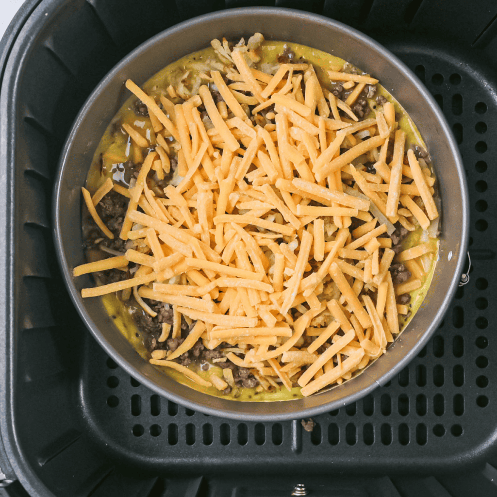  Pouring eggs into a nonstick pan in air fryer, topping with cooked ground beef, hashbrowns, and cheese, then cooking at 350°F.
