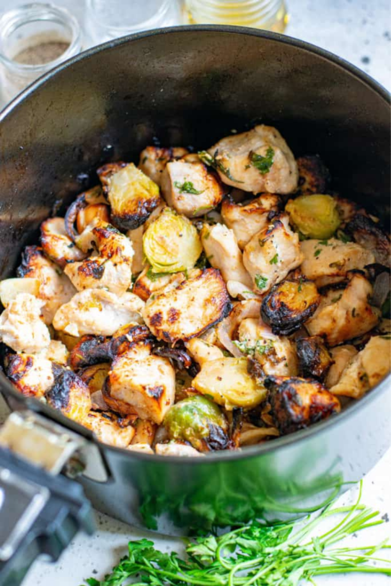 Air fryer basket filled with seasoned chicken and Brussels sprouts cooking, stirred halfway for even browning.