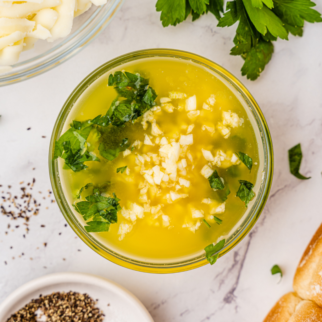 Hands mixing melted butter, minced garlic, and fresh chopped parsley in a small glass prep bowl for a slider glaze.