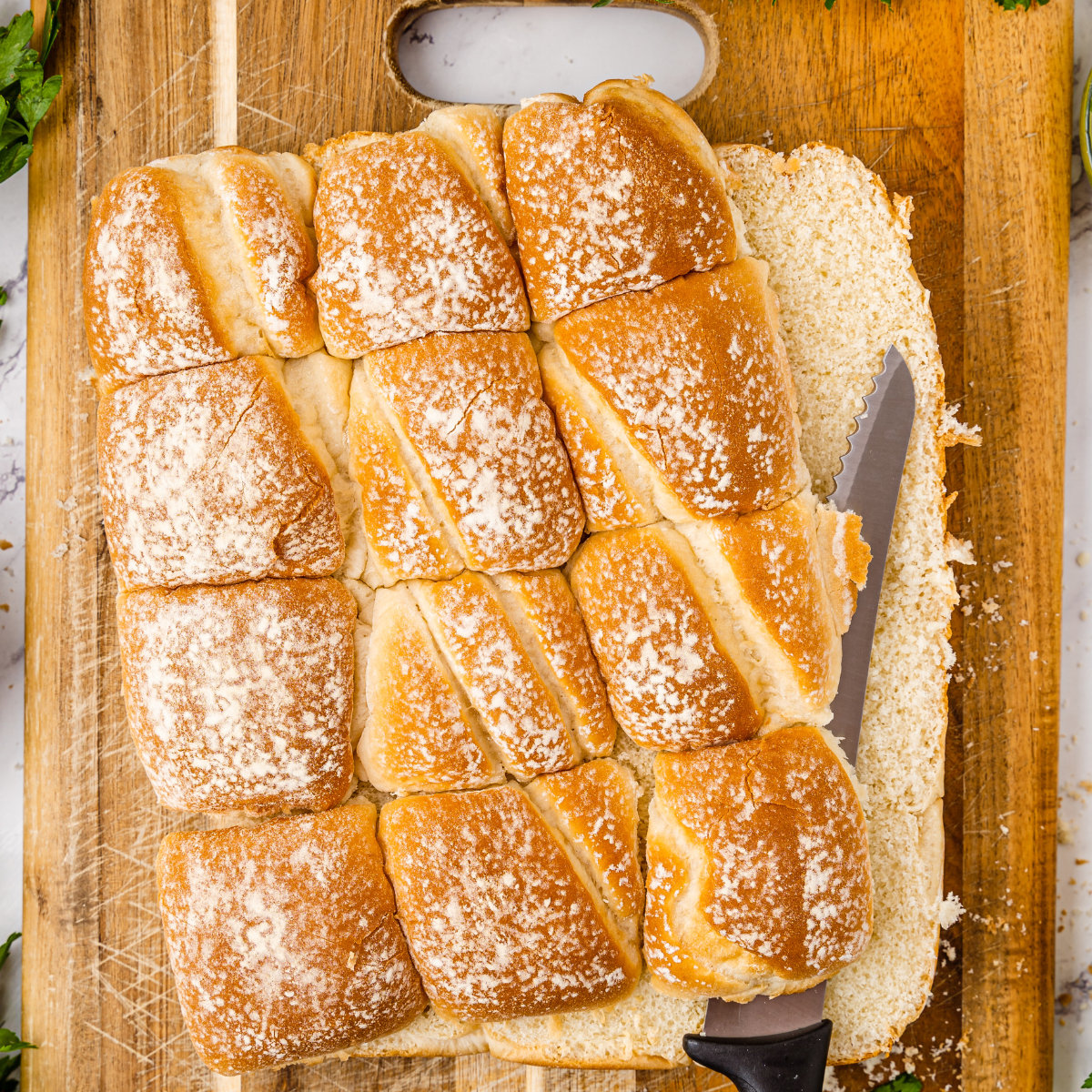 A long serrated knife slicing a full pack of slider rolls horizontally on a wooden cutting board to create top and bottom halves.