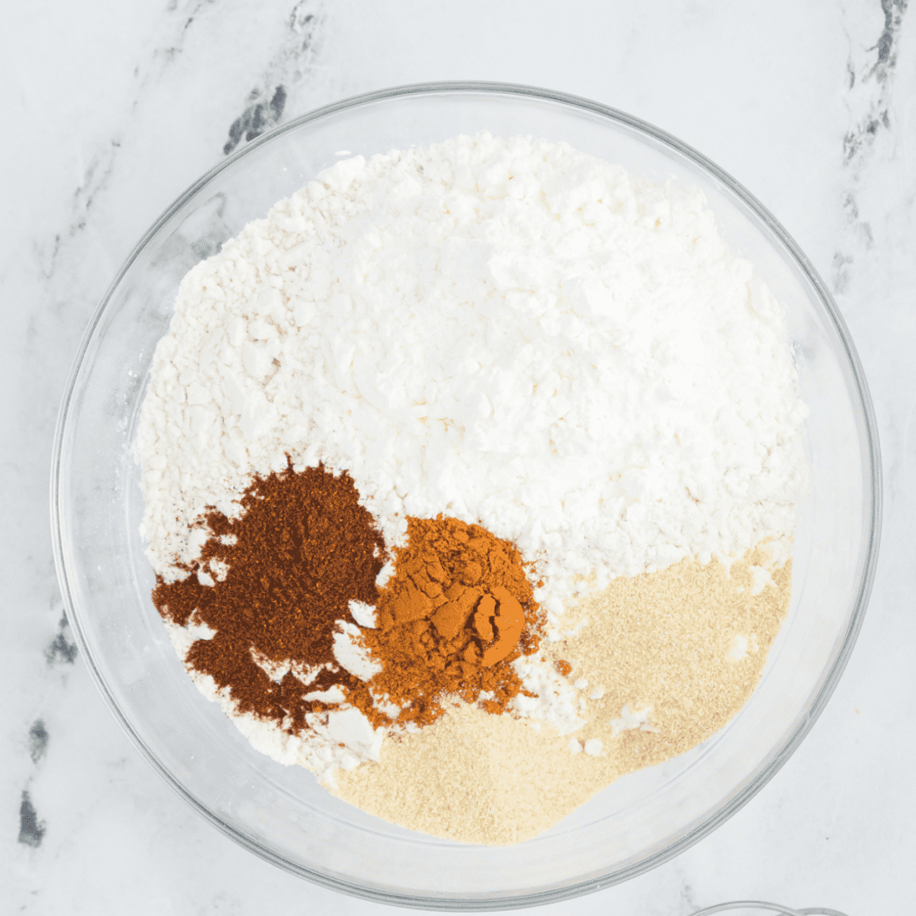 A bowl with a whisk combining all-purpose flour, cornstarch, cayenne pepper, paprika, onion powder, and garlic powder for a flavorful chicken coating mixture.