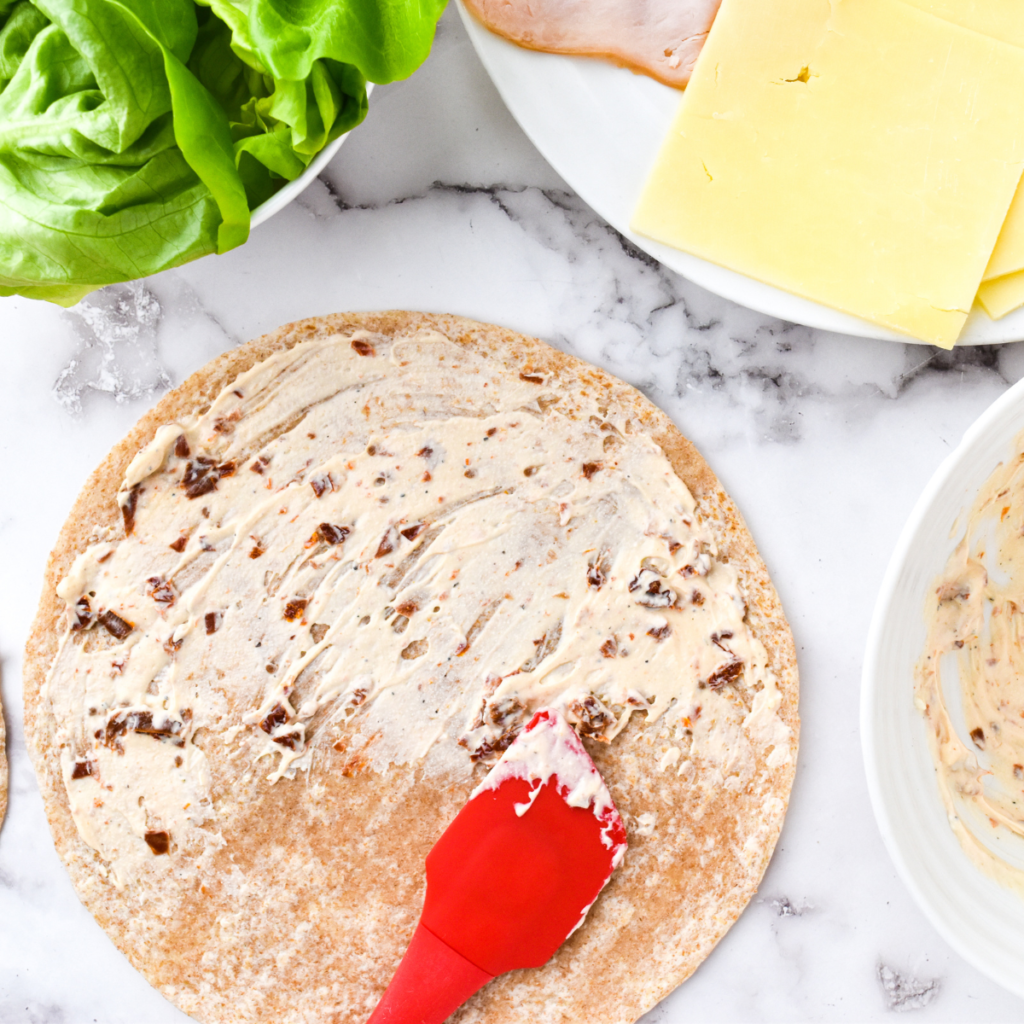 Flat tortilla on a cutting board spread with sun-dried tomato mixture, topped with cheese slices, turkey, and shredded lettuce, leaving an edge clear for rolling.
