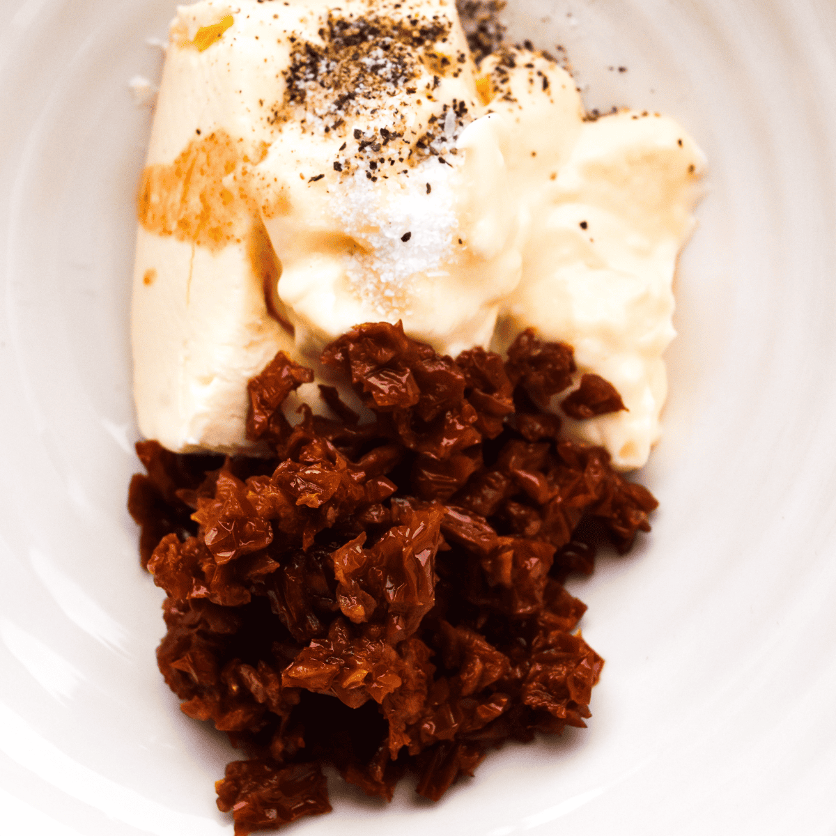 "Small bowl with a mixture of cream cheese, sun-dried tomatoes, mayonnaise, and Tabasco sauce being blended, seasoned with salt and pepper."