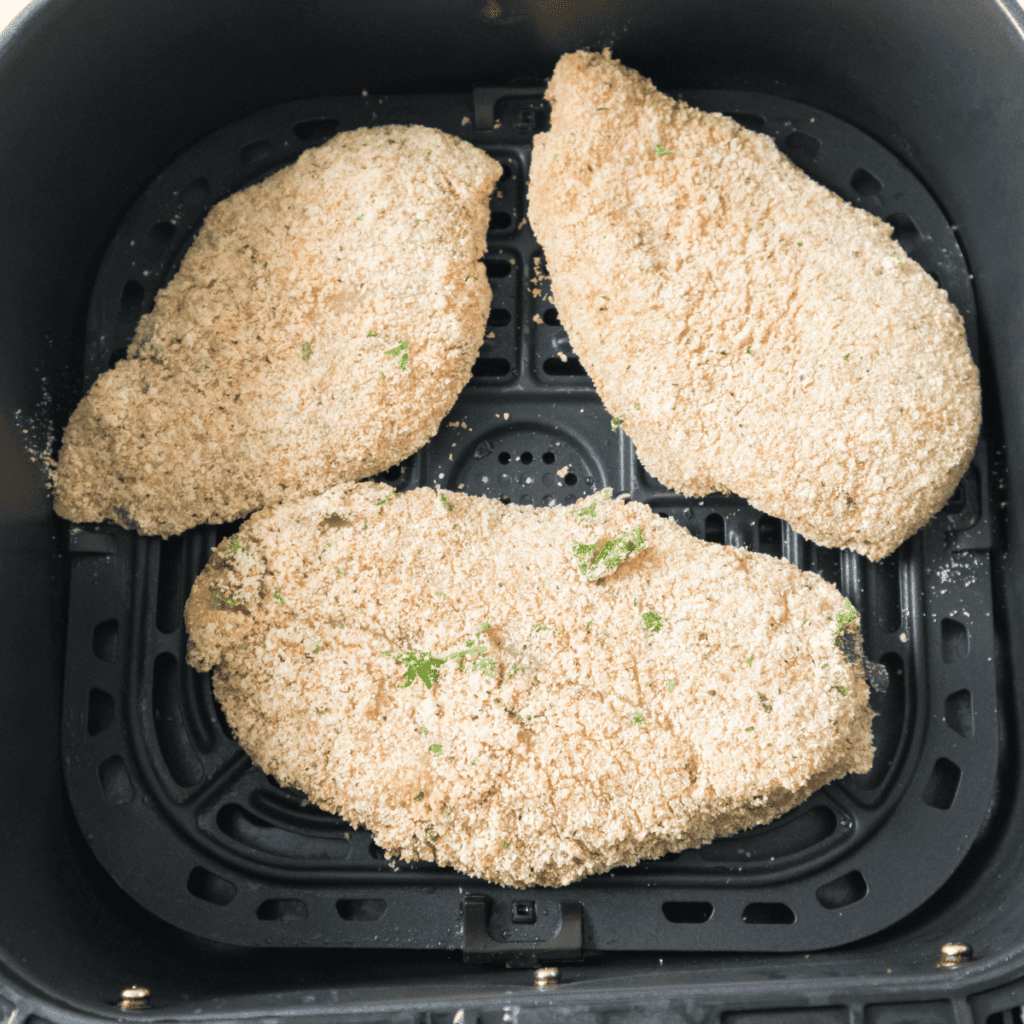 Air fryer basket being preheated and lightly sprayed with oil, alongside seasoned chicken breasts cooking at 375°F until golden and fully cooked, flipped halfway through.