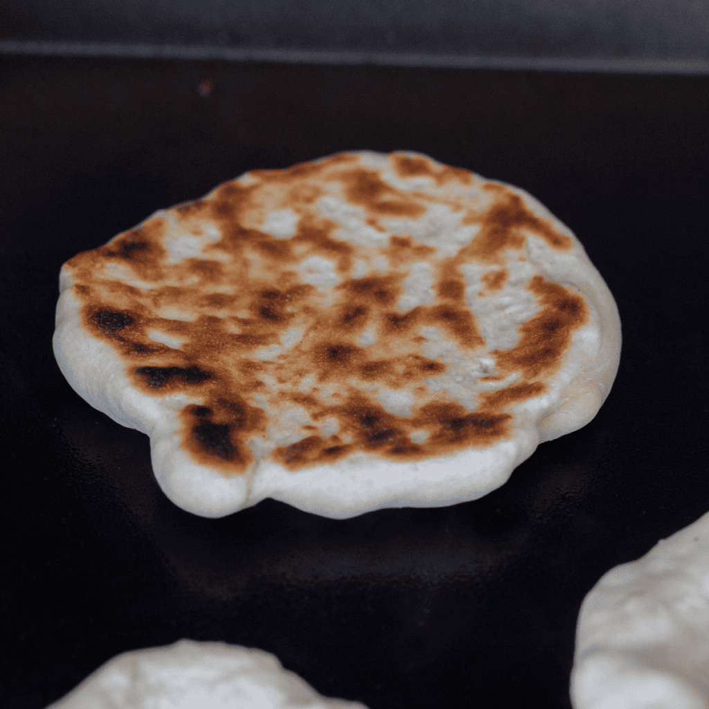 Preheated and oiled Blackstone griddle with naan dough cooking until golden brown on both sides.