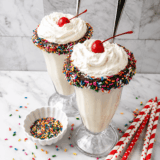Two Ninja Creami vanilla milkshakes on a table topped with whipped cream, sprinkles, and cherries