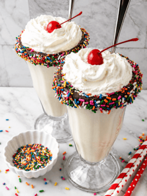 Two Ninja Creami vanilla milkshakes on a table topped with whipped cream, sprinkles, and cherries