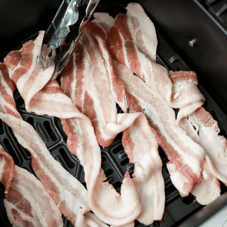 Bacon in the air fryer basket, with tongs on the side.