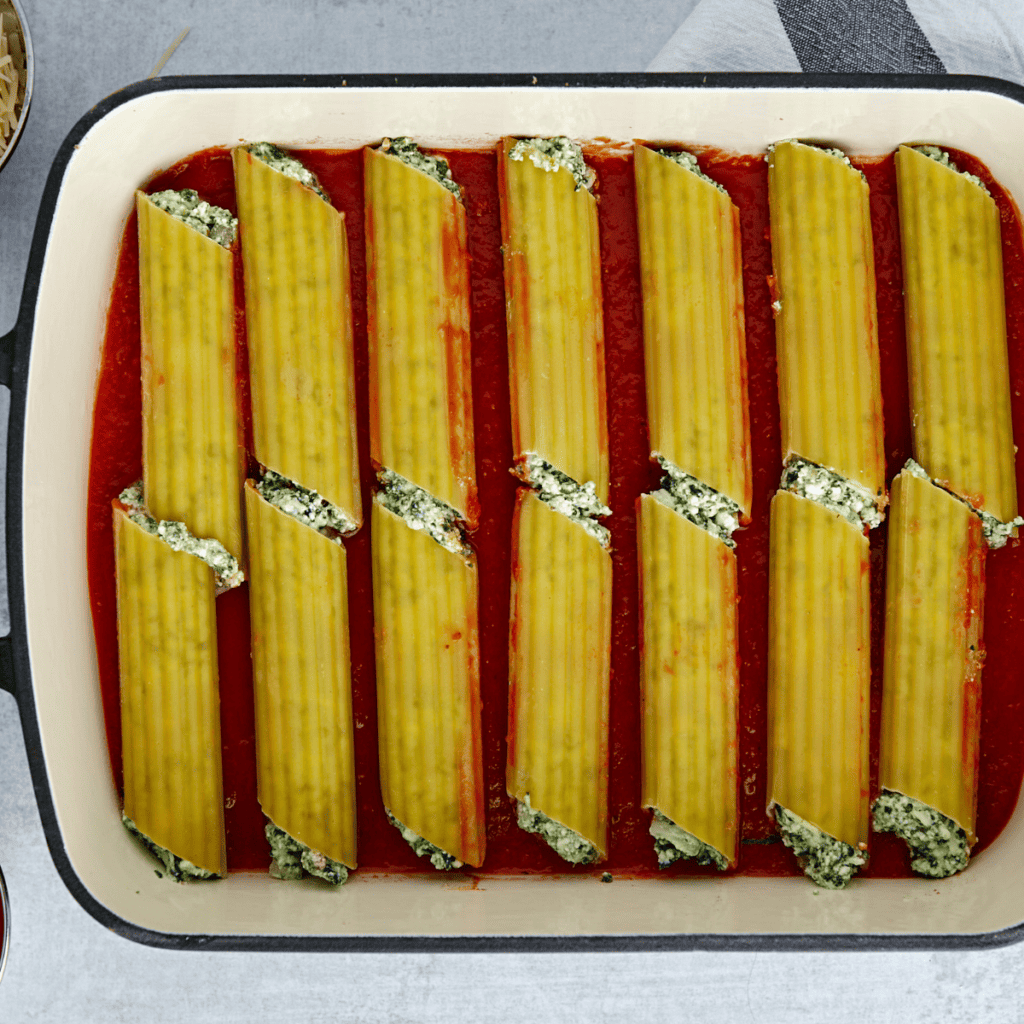Layering marinara sauce in a baking dish and placing stuffed manicotti inside, topped with remaining sauce