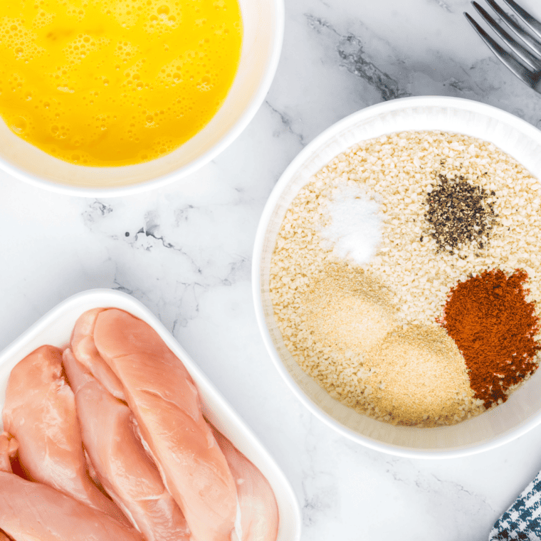 Chicken breasts sliced into strips being coated in a seasoned flour mixture in a shallow dish.