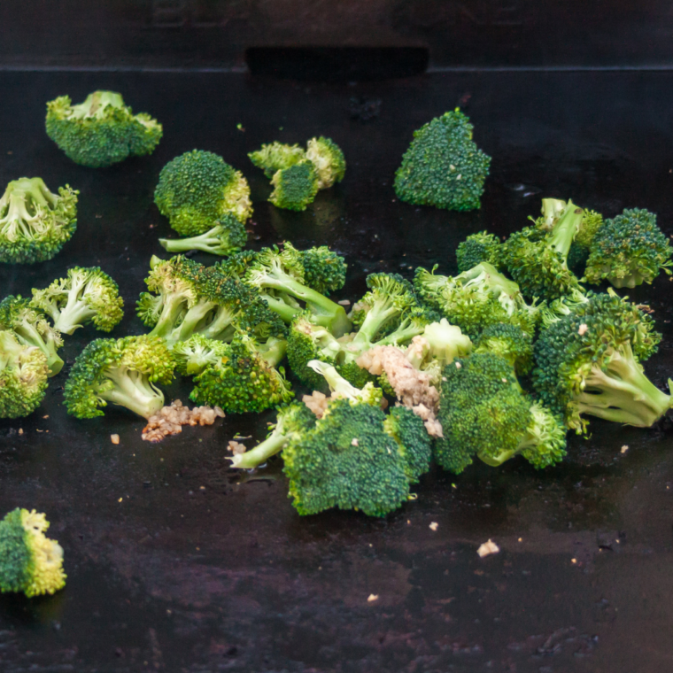 Fresh broccoli stir-frying on a Blackstone griddle until bright green and tender-crisp with light steam from a basting dome.