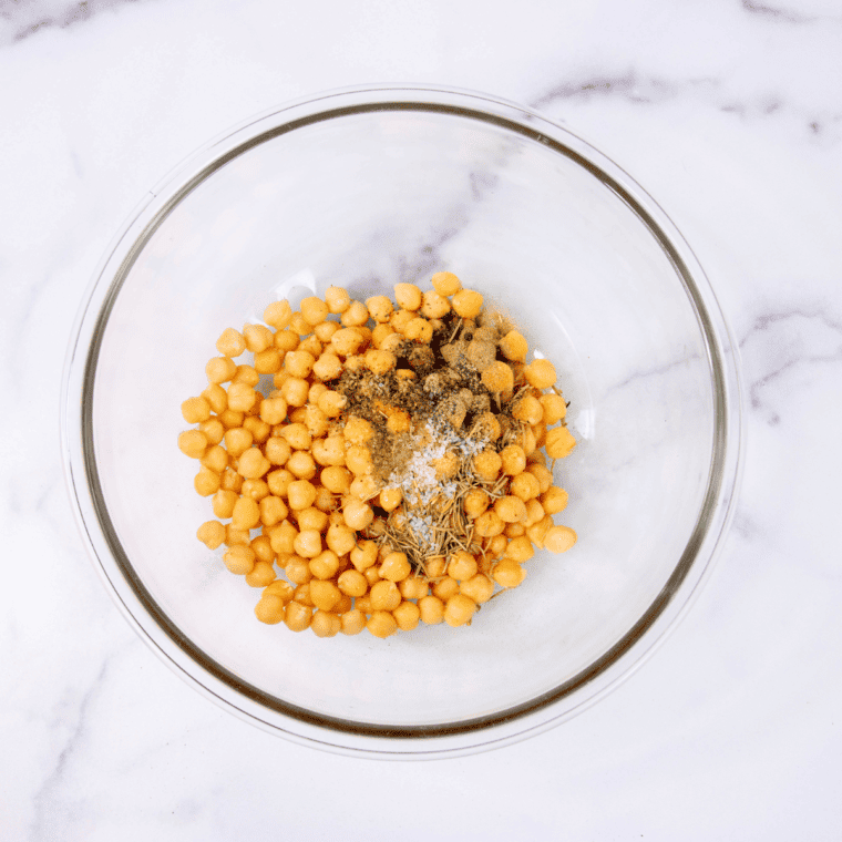 Chickpeas in a bowl being tossed with olive oil, garlic powder, dried rosemary, thyme, oregano, sea salt, and black pepper.