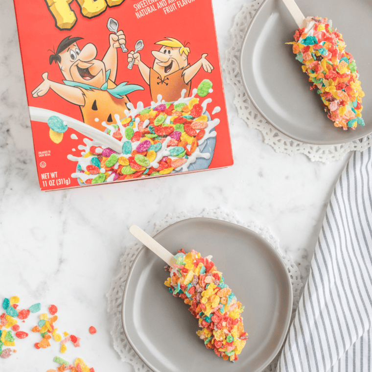 Close-up of Fruity Pebbles Banana Pops with chocolate coating and colorful cereal topping on a wooden serving tray.