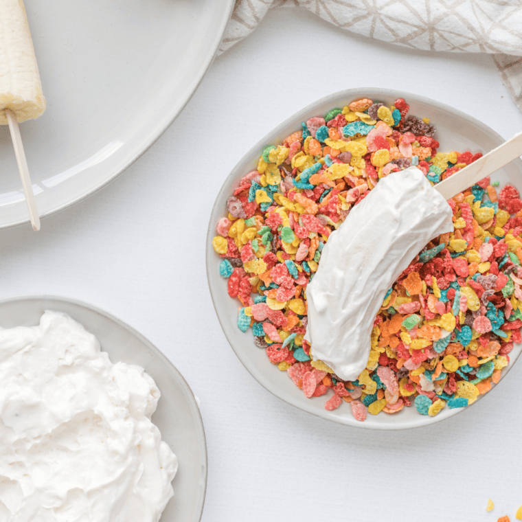 Banana halves coated in strawberry yogurt mixture being rolled in Fruity Pebbles cereal on a shallow dish.