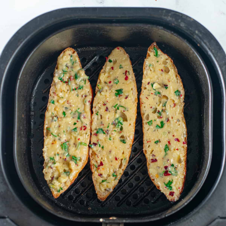 Buttered baguette slices arranged butter-side up in a single layer inside an air fryer basket, ready to cook
