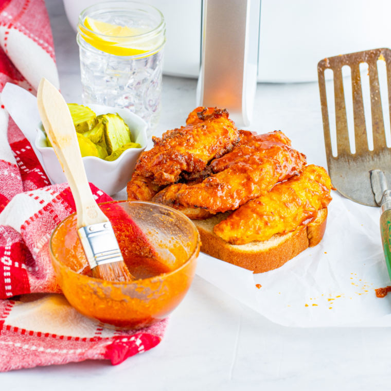 Overhead flat lay of air fryer Nashville hot chicken tenders on parchment paper with white bread, pickles, and a small bowl of cayenne hot oil