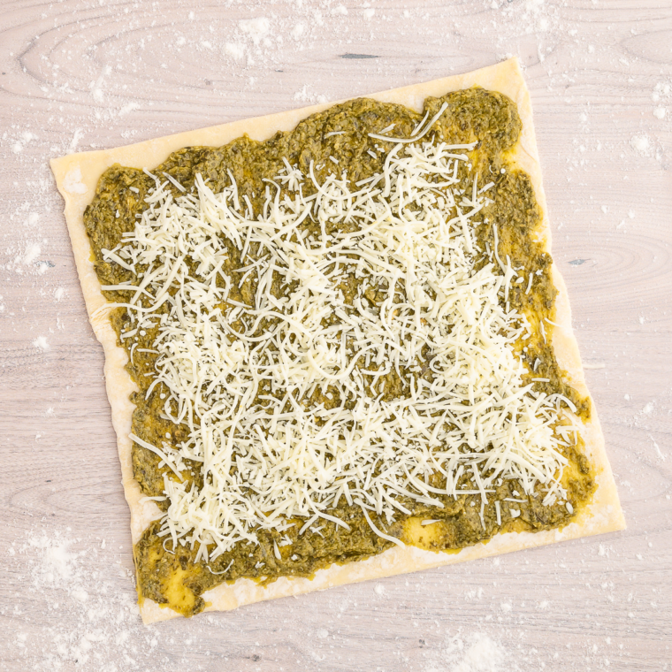 Adding cheese to the pesto-covered puff pastry before rolling it up."
