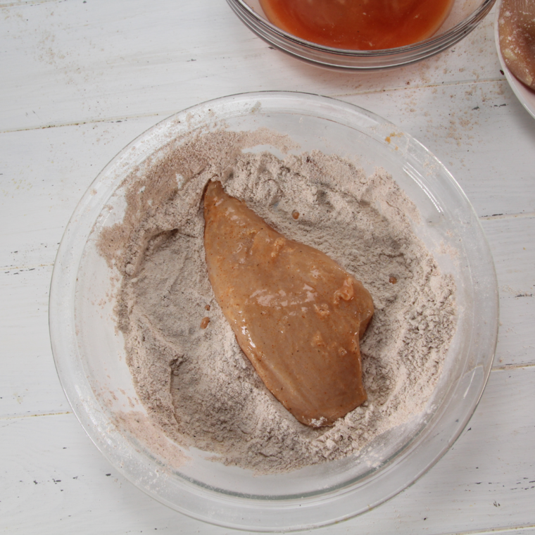 Chicken breast being coated in a seasoned flour mixture, dipped into hot sauce, and coated again with flour for a crispy breading.