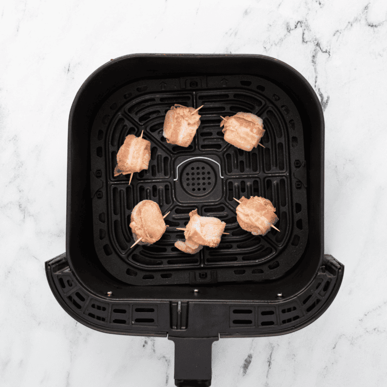 Frozen bacon-wrapped scallops arranged in a single layer in the air fryer basket for even cooking.