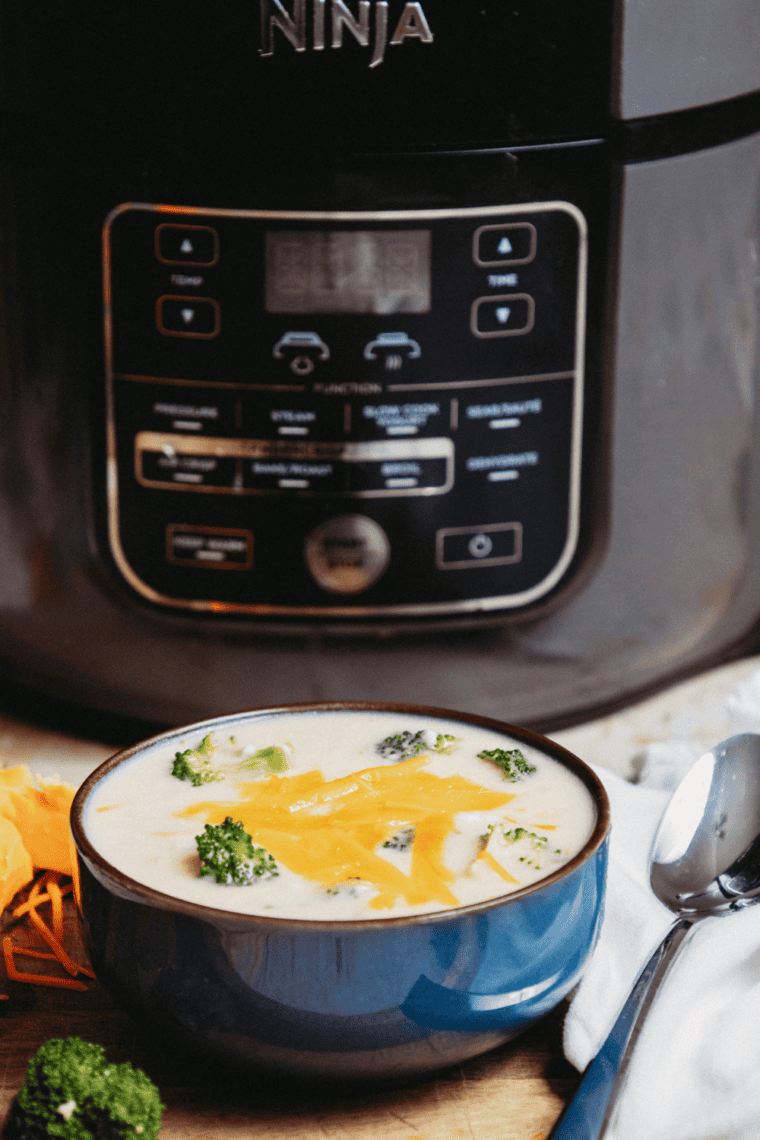 A bowl of creamy broccoli cheddar soup cooked in the Ninja Foodi, topped with shredded cheddar and broccoli florets.