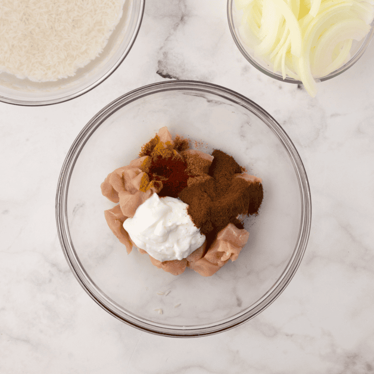 Soaked basmati rice in a bowl and marinated chicken pieces coated with yogurt and spices, resting before cooking.