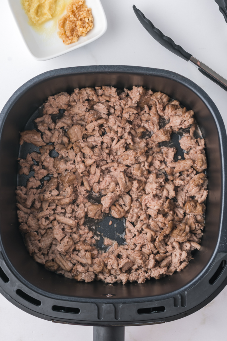 An air fryer basket with browned ground meat, seasoned and mixed with vegetables in preparation for the egg roll bowl.
