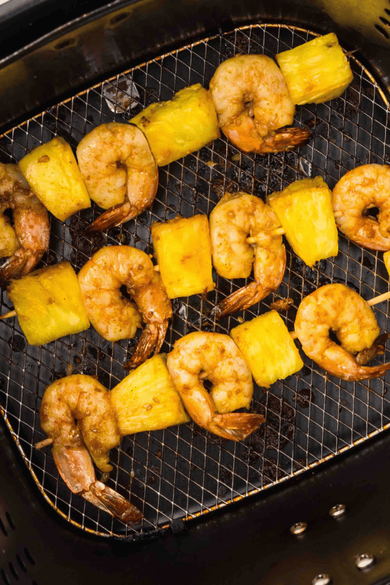 Shrimp and pineapple skewers cooking in air fryer basket brushed with teriyaki sauce.