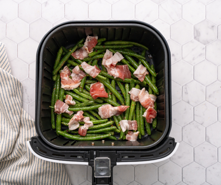 Seasoned green beans and diced bacon arranged in a single layer inside an air fryer basket.