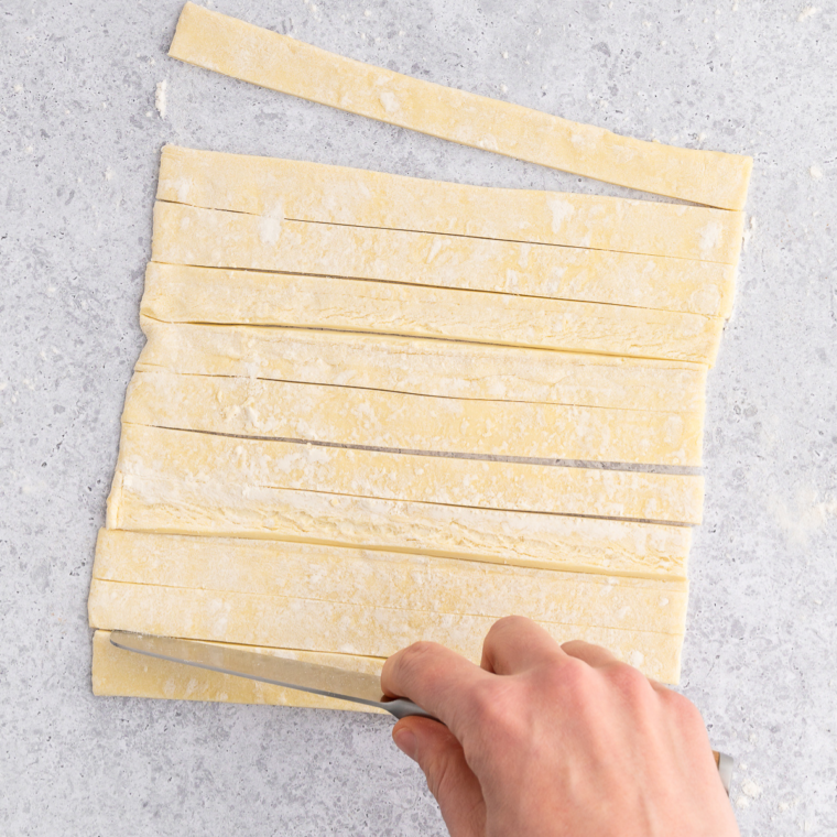 Puff pastry sheet being cut into ½ inch wide strips with a pizza cutter on a cutting board.