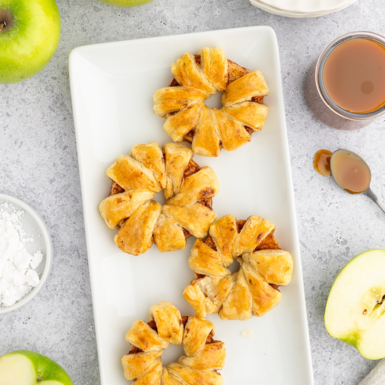 Golden brown Air Fryer Puff Pastry Apple Rings arranged on a white plate, garnished with a sprinkle of powdered sugar and surrounded by fresh apple slices.