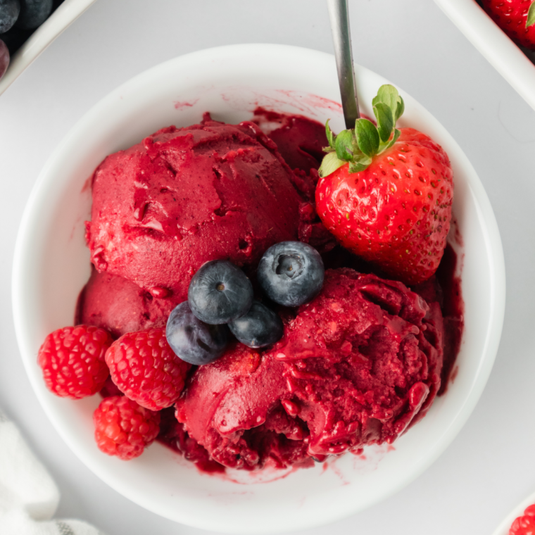 Close-up of mixed berry sorbet in a white bowl, garnished with fresh fruit and served with a spoon.
