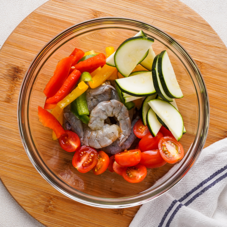 Cut vegetables and shrimp in a glass bowl, on a cutting board.