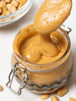 A jar of smooth, homemade peanut butter made in the air fryer, with peanuts scattered around on a wooden surface.